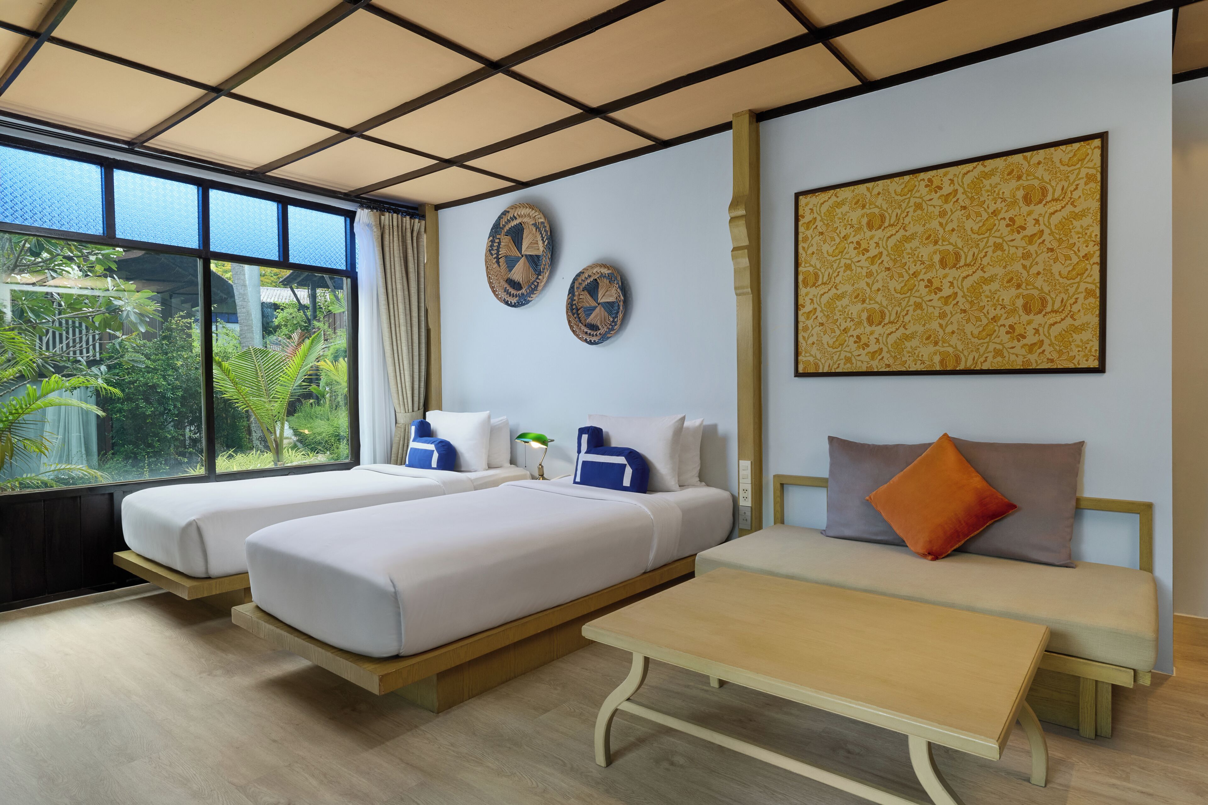 A hotel room with two beds, a sofa, a table, and large windows with a view of greenery.