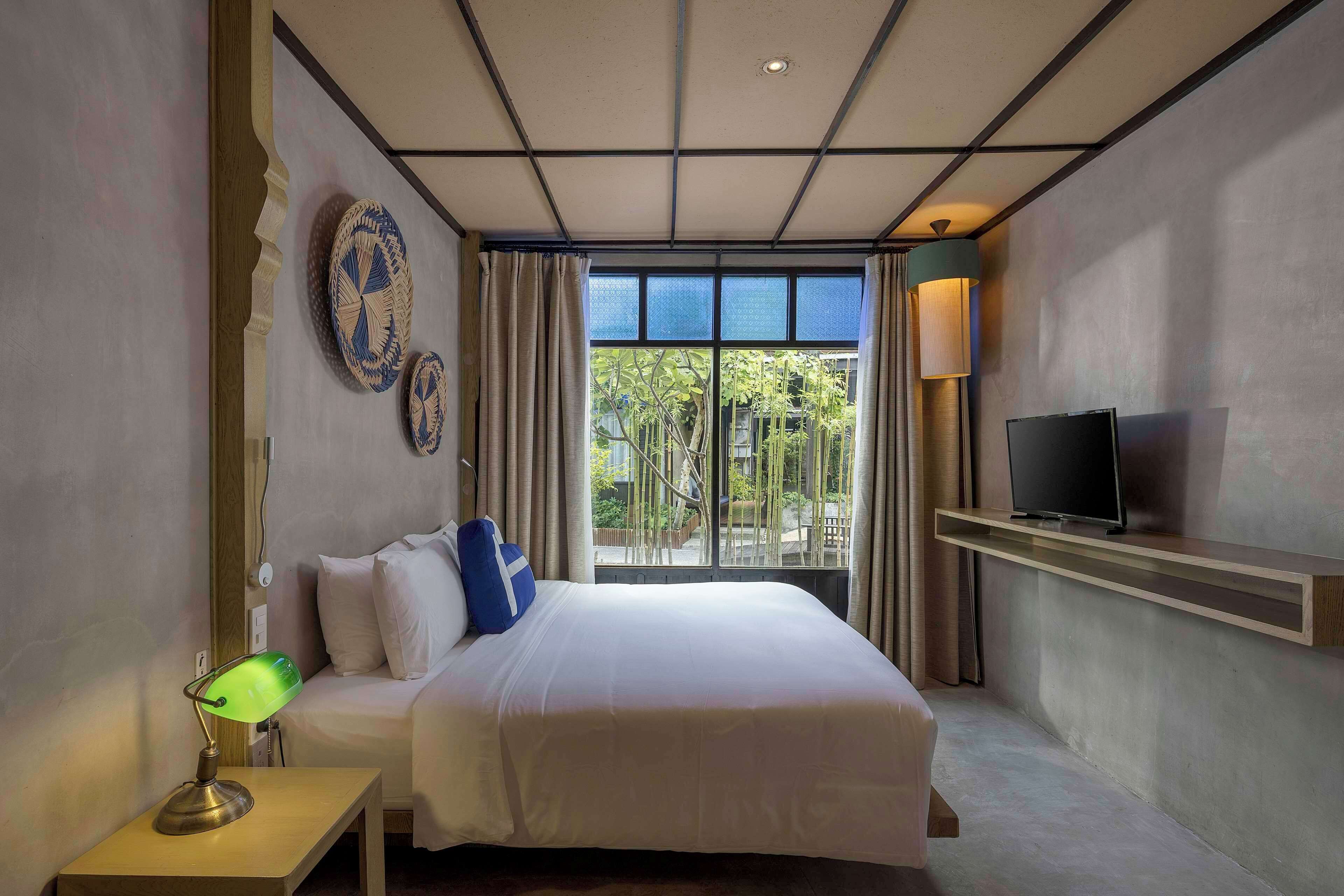 A hotel room with a bed, a TV on a shelf, a desk with a lamp, and a window with a view of greenery.