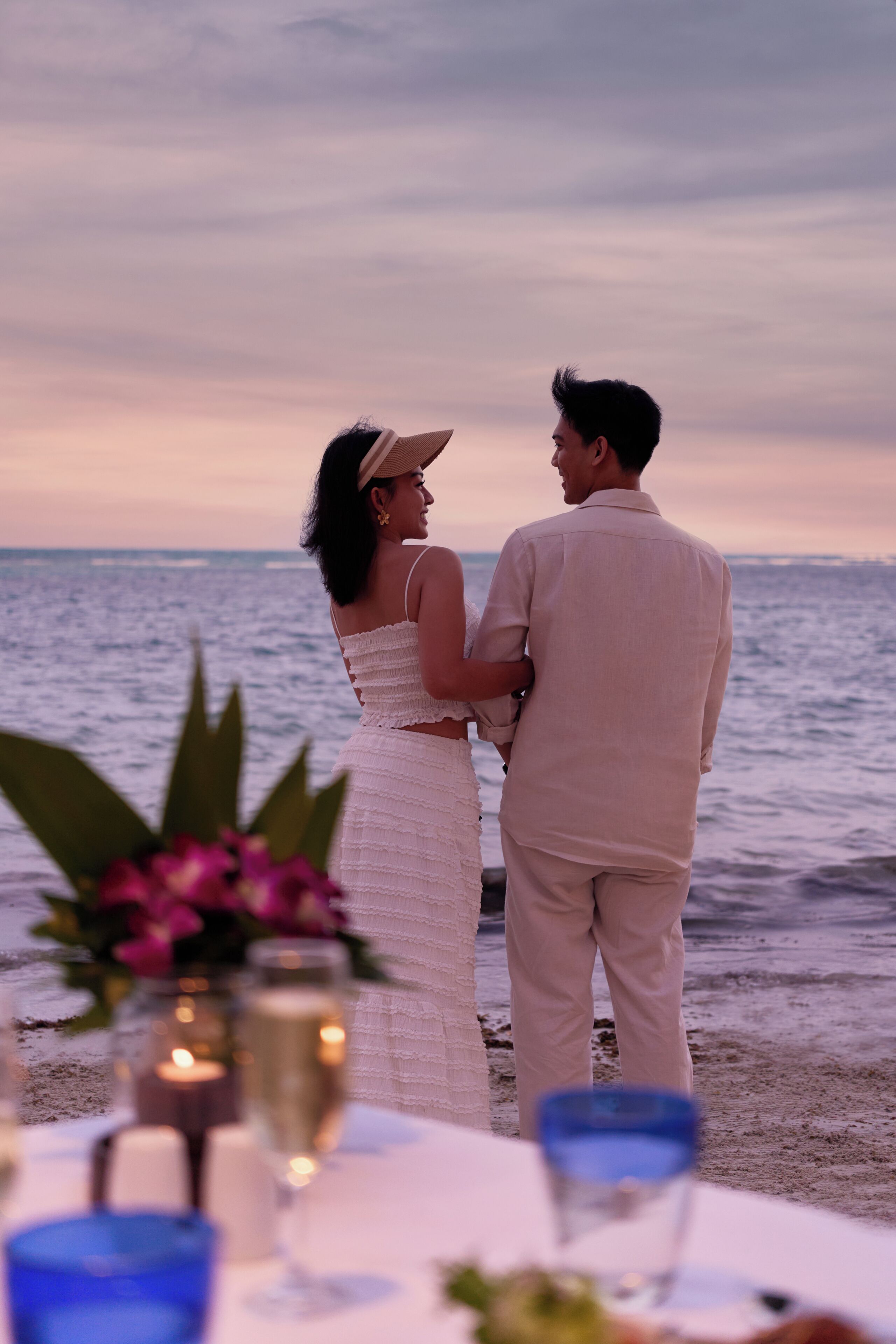Una pareja de pie junto a una mesa, con vista al mar, una vela y un vaso de agua.