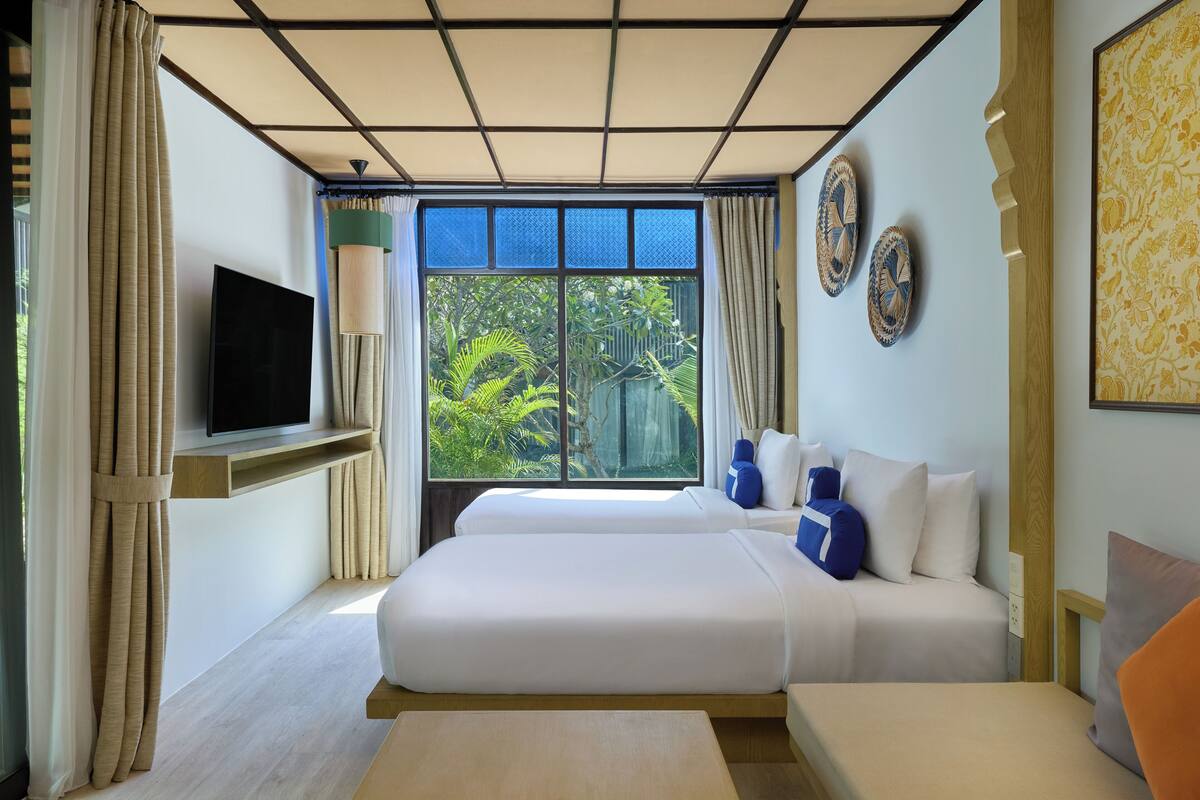 A hotel room with two beds, a TV, a window with a view of greenery, and decorative plates on the wall.