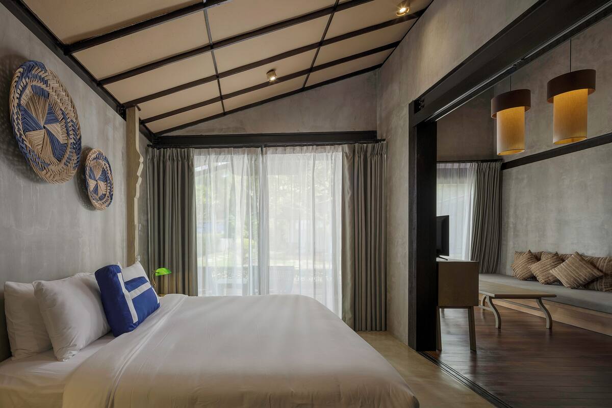 A modern hotel room with a large bed, a seating area with cushions, and a view through a glass door.