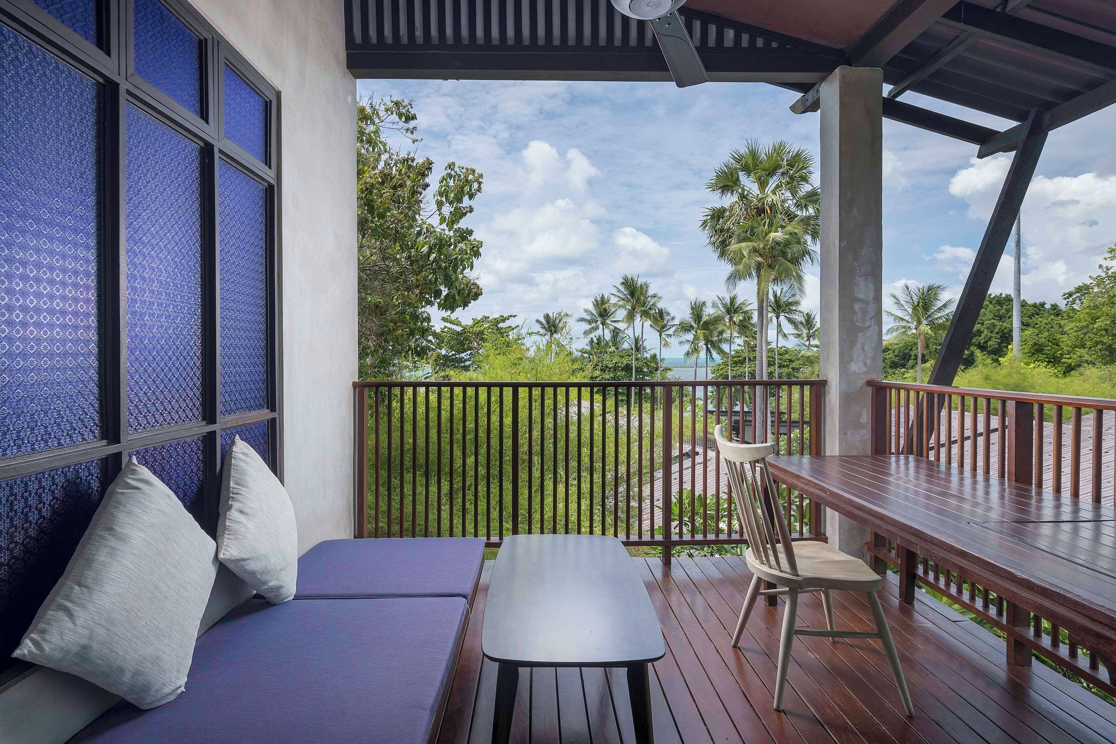 A balcony with a wooden table and chairs, a cushioned bench, and a view of palm trees and a beach.