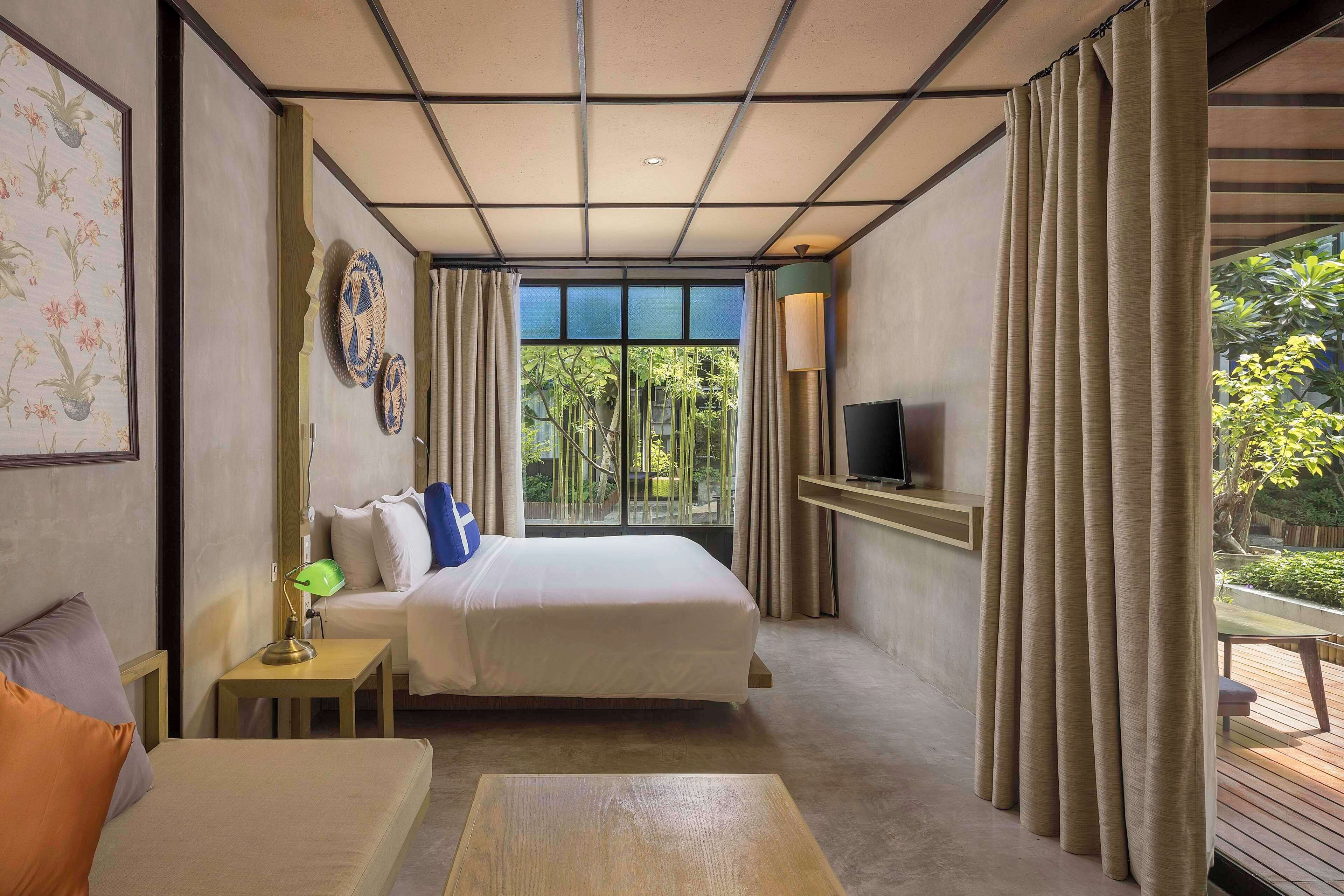 A hotel room with a bed, a desk, a lamp, a TV on a shelf, and a view of greenery outside.