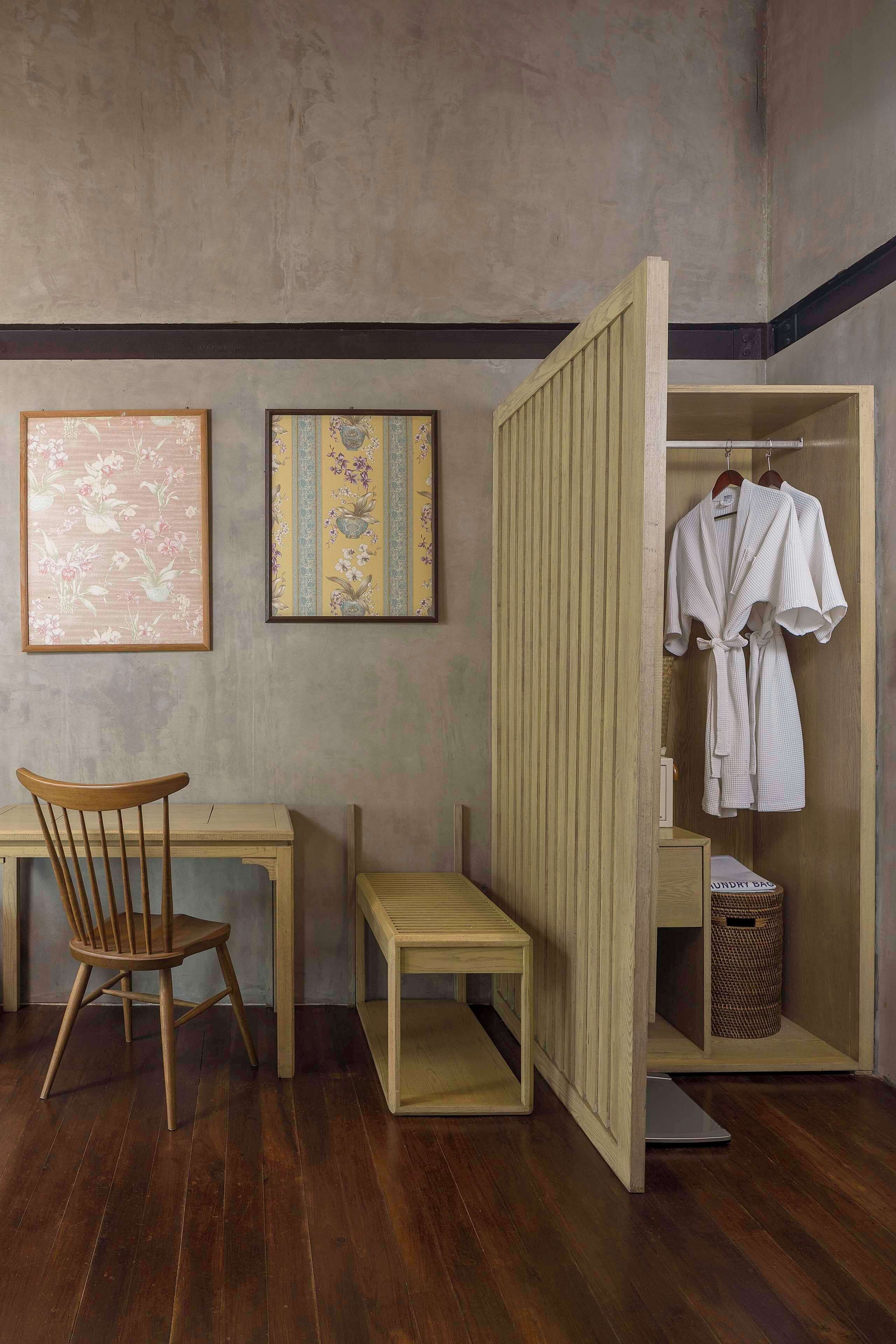 A room with a wooden desk, a chair, a small table, and a clothing storage unit with hanging robes.