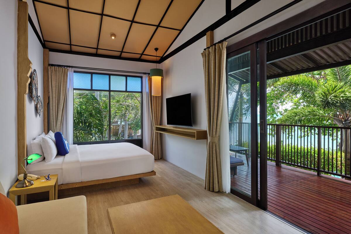 A modern hotel room with a bed, TV, and balcony with a view of greenery.