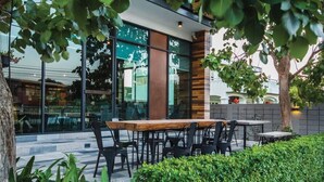An outdoor seating area with a wooden table and chairs, surrounded by greenery and a modern building facade. - Space 59 Ratchaburi (Ratchaburi)
