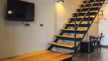 Una escalera moderna con escalones de madera y barandillas metálicas, un televisor de pantalla plana montado en la pared, y una pequeña mesa con sillas al fondo.