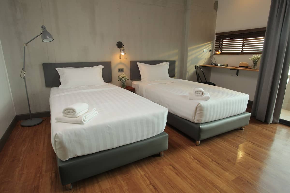 A hotel room with two beds, a standing lamp, a nightstand, a shelf with decorative items, and a window with blinds.
