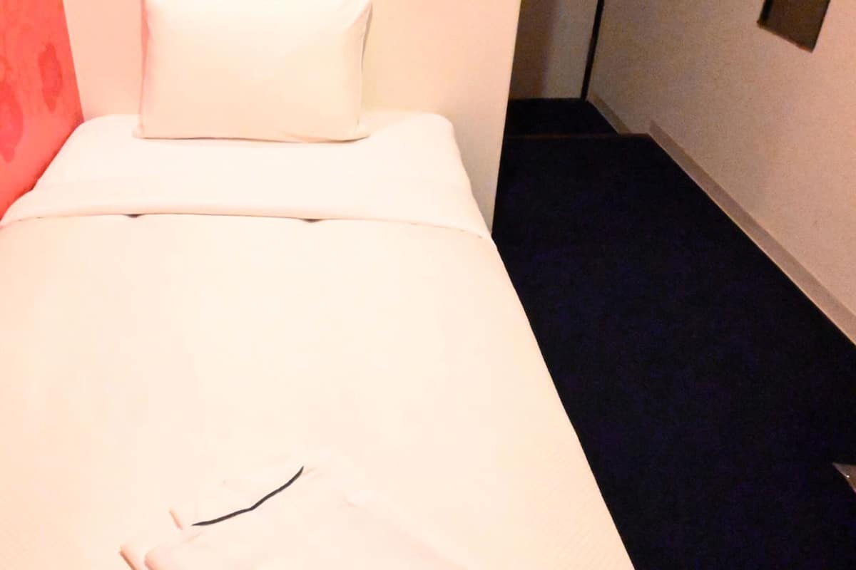 Un letto singolo con materasso e cuscino bianchi, un piccolo oggetto nero sul letto e una parete rossa con motivi a sinistra.