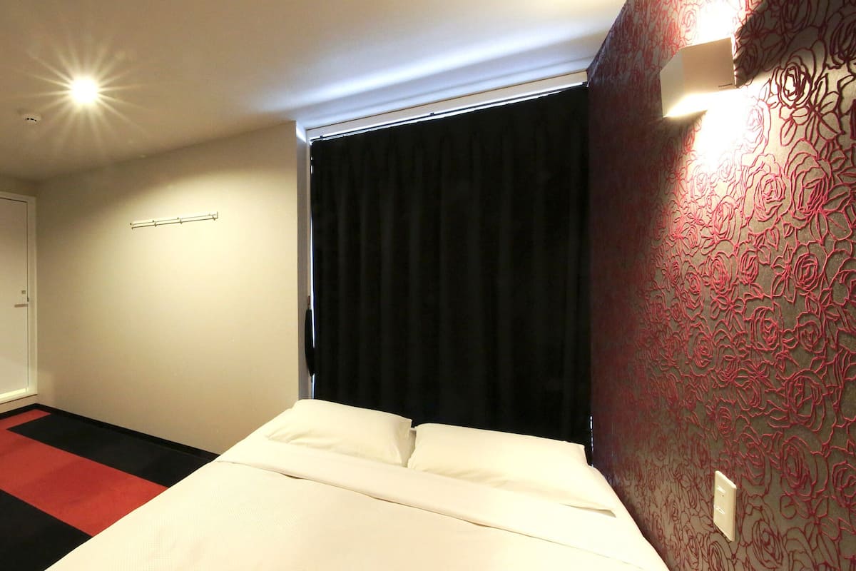 Una camera d'hotel con un letto, un comodino, uno specchio, una piccola pianta e una finestra con tenda nera.