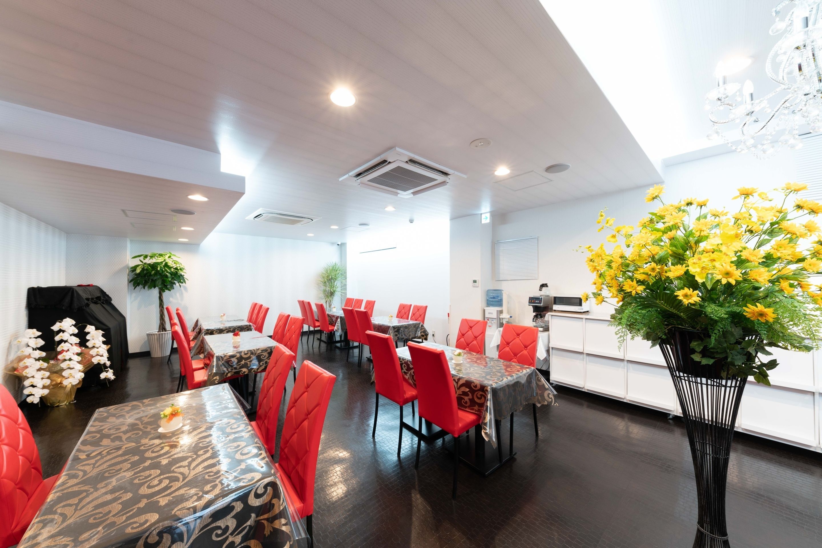 Un comedor de hotel con sillas rojas, mesas con manteles estampados y un jarrón grande con flores amarillas.