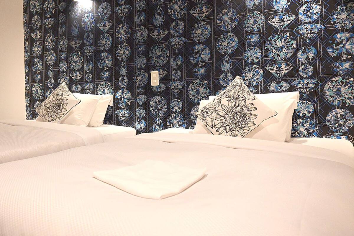 Camera d'albergo con due letti, ognuno dotato di copripiumino bianco e cuscini abbinati. Sullo sfondo, carta da parati con motivi floreali in blu e nero.
