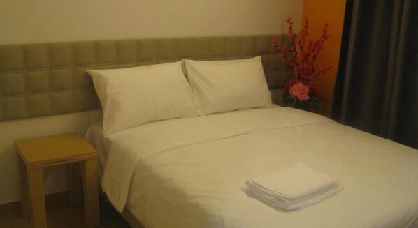 A neatly made bed with white linens, a small wooden nightstand, and a vase with pink flowers.