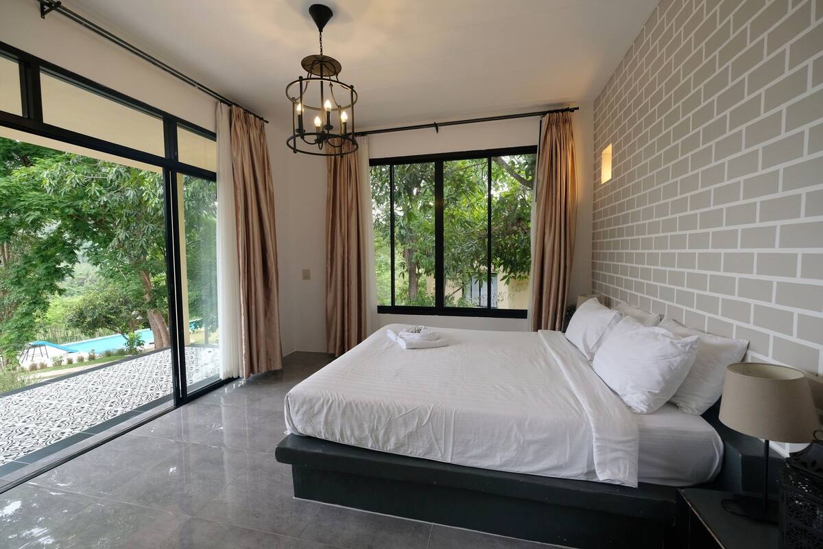 A modern bedroom with a large bed, a chandelier, and a view of greenery outside.