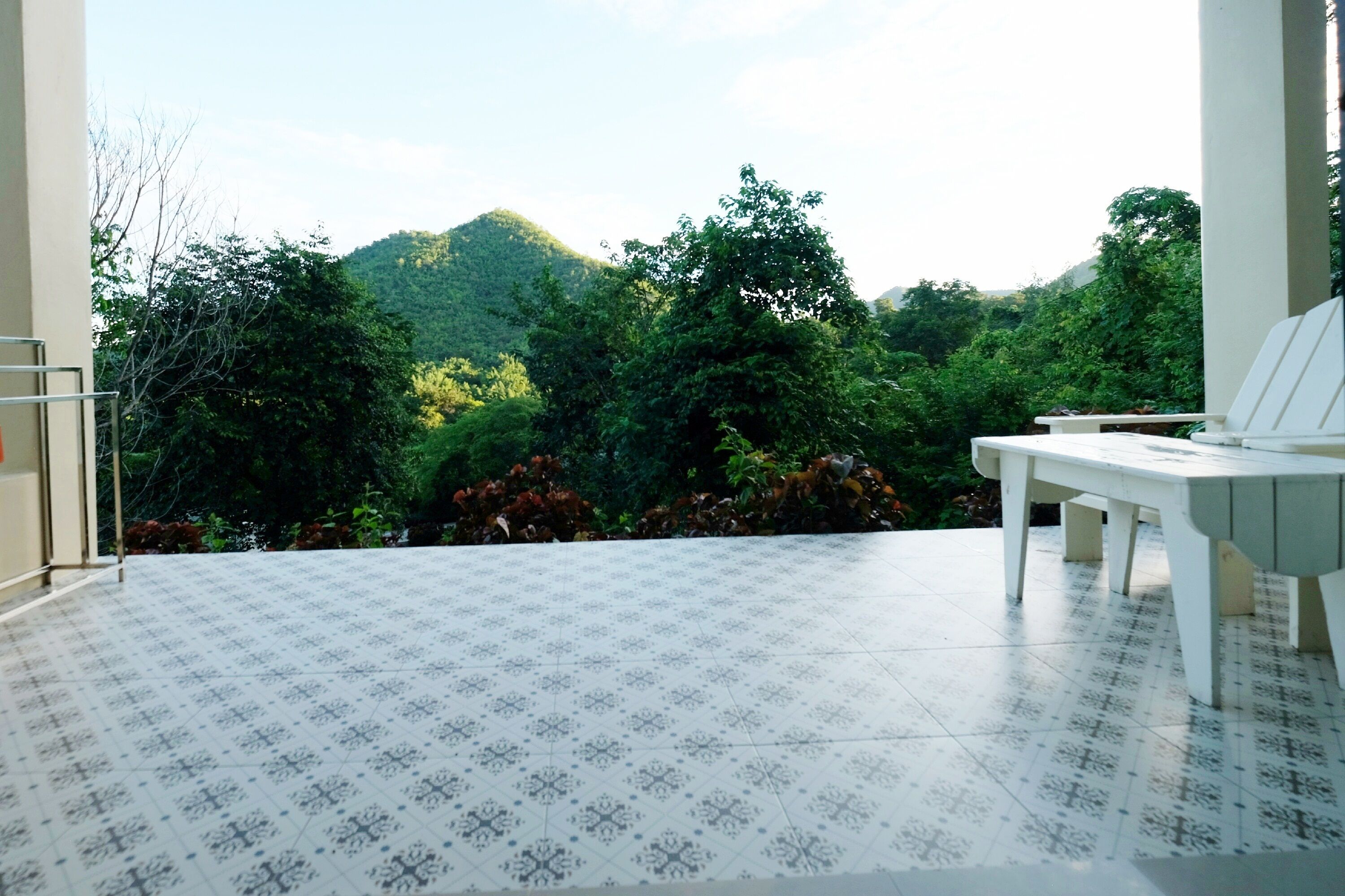 A balcony with a patterned floor and a white chair, overlooking a lush green landscape with trees and hills.