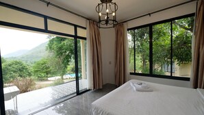 A hotel room with a large bed, a chandelier, and a view of greenery and mountains through large windows. - Hi - Scene Resort (Suan Phueng)