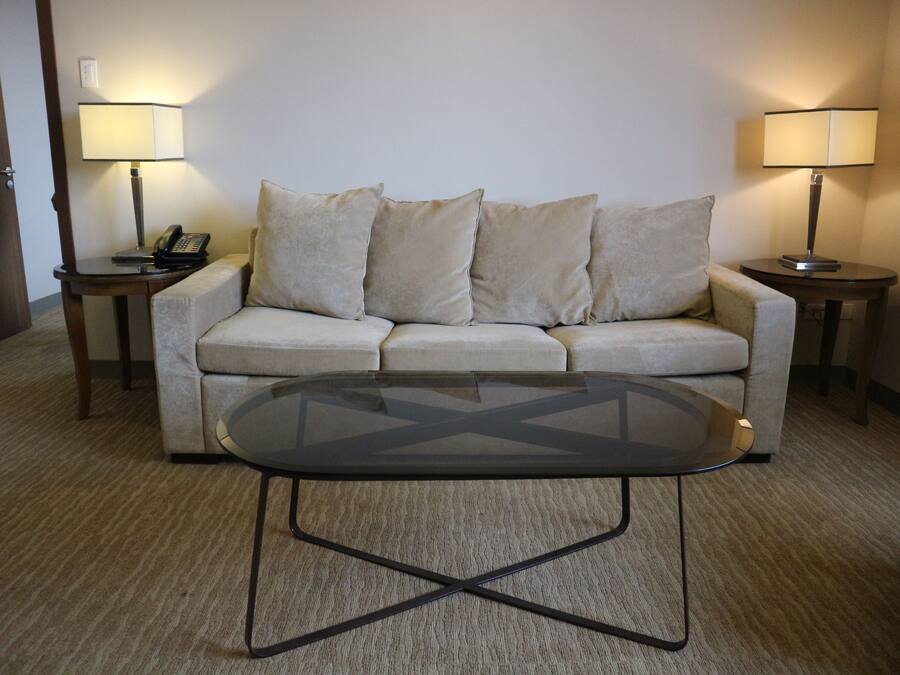 A hotel room with a beige sofa, two bedside tables with lamps, and a glass coffee table.