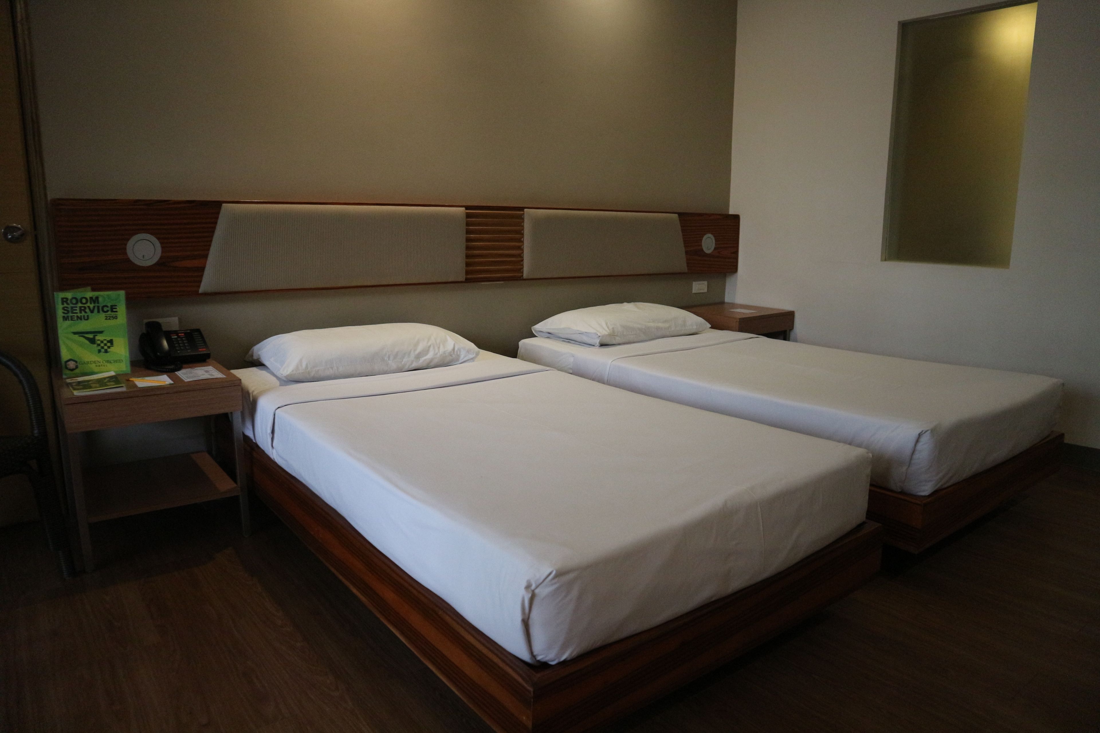 Une chambre d’hôtel avec deux lits simples, une tête de lit en bois, une table de chevet avec un téléphone et un luminaire fixé au mur.