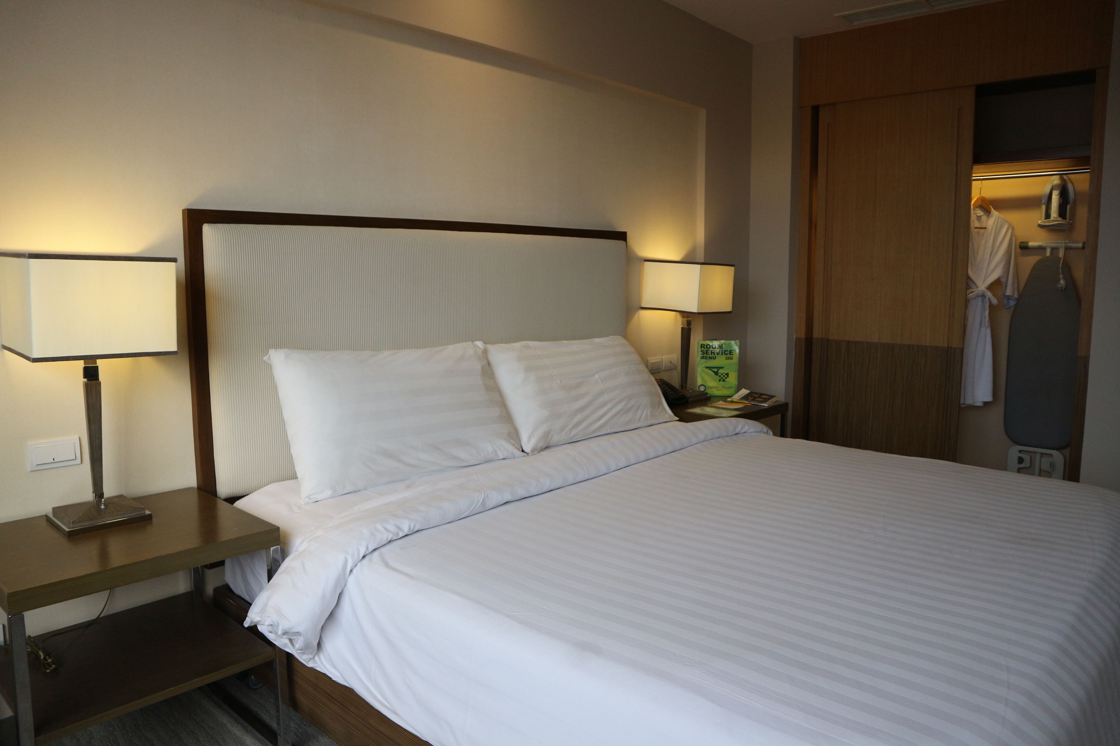 A hotel room with a bed, two bedside lamps, a nightstand, a robe, and an ironing board.
