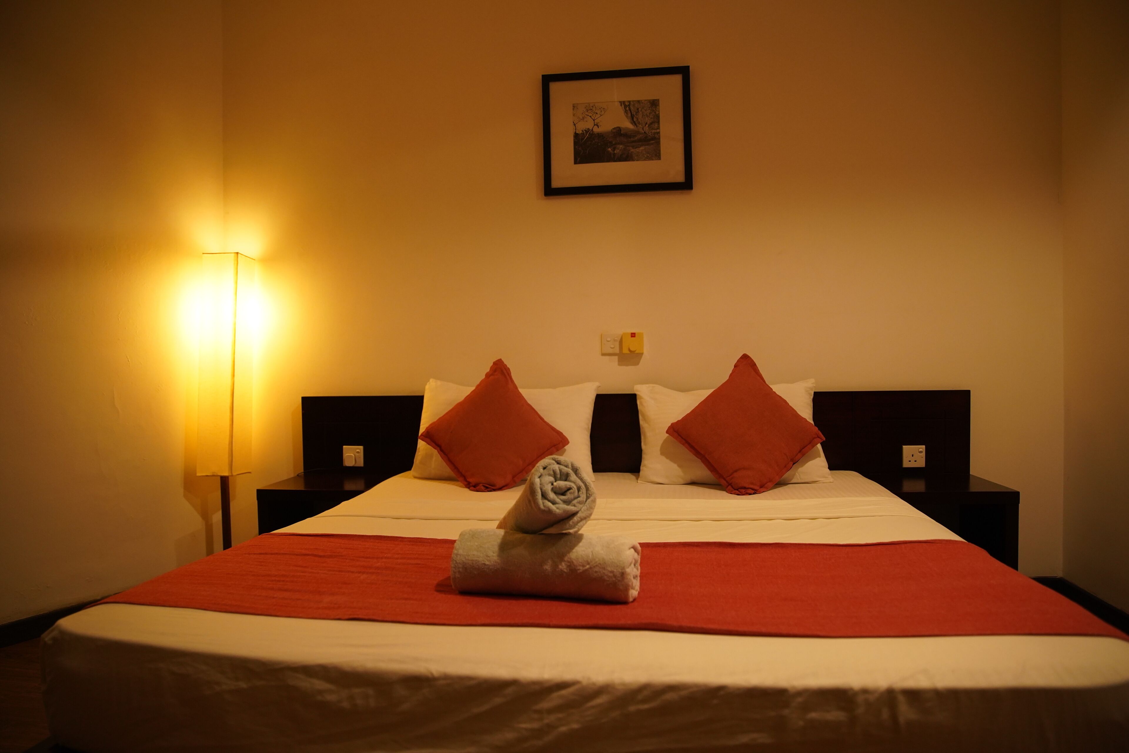Ein Hotelzimmer mit einem ordentlich bezogenen Bett, roten Zierkissen, einem gerahmten Bild an der Wand und einer Lampe auf dem Nachttisch.