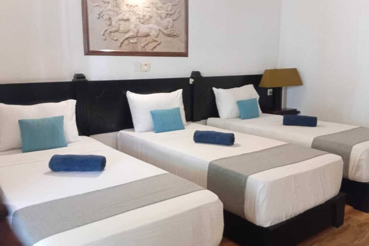 Habitación de hotel con cuatro camas individuales, cada una con colchón blanco y almohadas azules. Hay una obra de arte enmarcada sobre las camas y una lámpara en la mesita de noche.