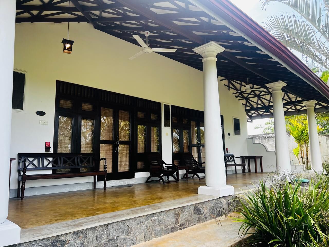 A covered outdoor seating area with wooden benches and chairs, a ceiling fan, and a palm tree in the background.