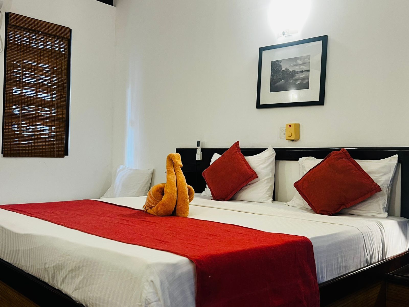Een hotelkamer met een bed, rode kussens, een lijst met een foto aan de muur en een raam met zonwering.