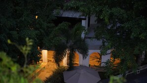 An outdoor patio area with a large umbrella, surrounded by lush greenery and architectural features including a stone wall and a small building with a tiled roof. - Fort Edge Retreat (Unawatuna)