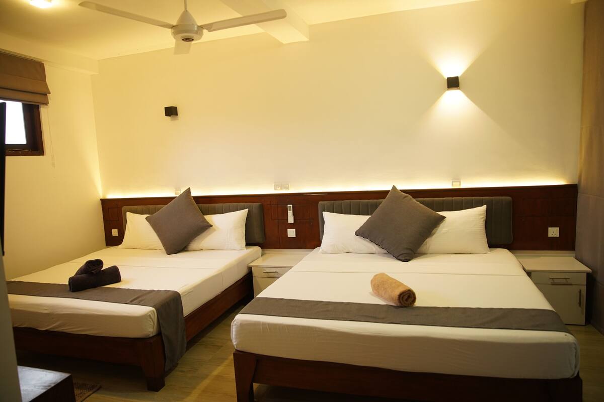 Habitación de hotel con dos camas individuales, ventilador de techo, lámparas de noche, mesitas de noche y una ventana con cortinas.