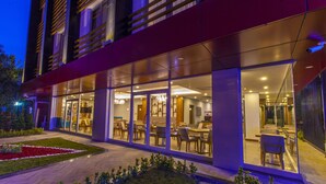 Fachada moderna de un hotel con puertas de vidrio, asientos exteriores e interior bien iluminado.