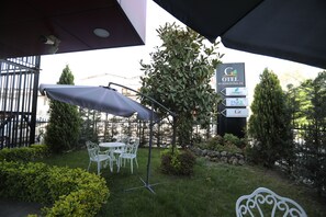 Un área de estar al aire libre con sillas de metal blanco y una mesa bajo un paraguas grande, rodeada de follaje y un letrero que dice 'GOTEL BUSINESS PALAS'.