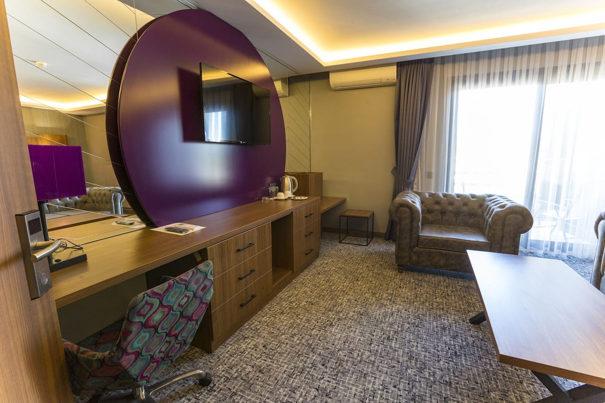 A hotel room with a wooden desk, a purple accent wall, a television, a sofa, and a large window.
