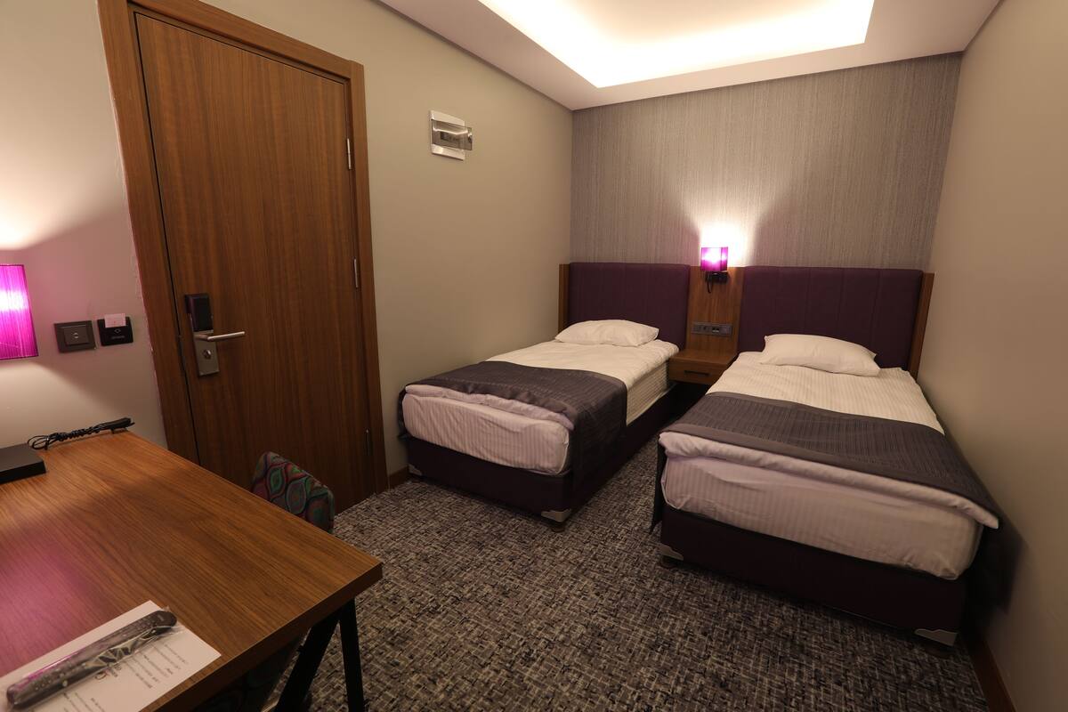 A hotel room with two single beds, a wooden desk, a chair, a small table, a door, and a wall-mounted light.