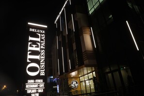 Un edificio con un letrero que dice "OTEL BUSINESS PALAS DELUXE" y que anuncia servicios como sauna, gimnasio, restaurante, cafetería, reuniones y salón de eventos.