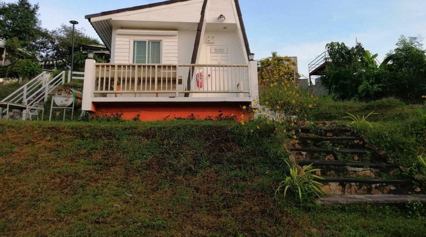 Un edificio blanco con balcón y base roja, rodeado de vegetación y escaleras.