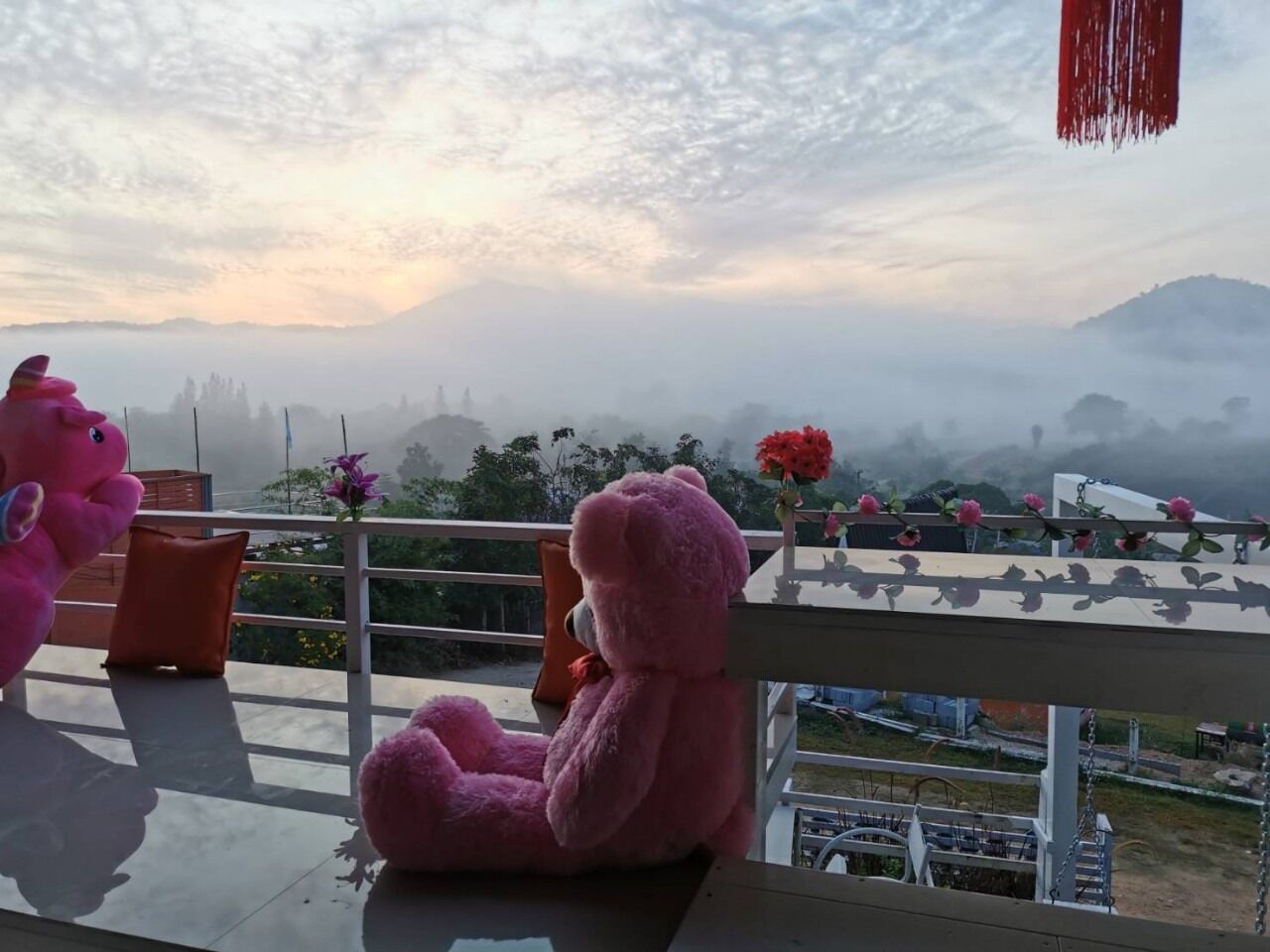 A balcony with a pink teddy bear, a table with a vase of flowers, and a view of a misty landscape.