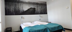 A hotel room with a bed, a nightstand, a wall-mounted light, and a large painting of a dock and water.