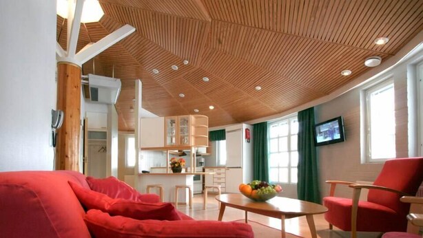 A modern living room with a red sofa, a wooden ceiling, and a kitchen in the background. - Hotelli Pyöreä Torni (Savonlinna)
