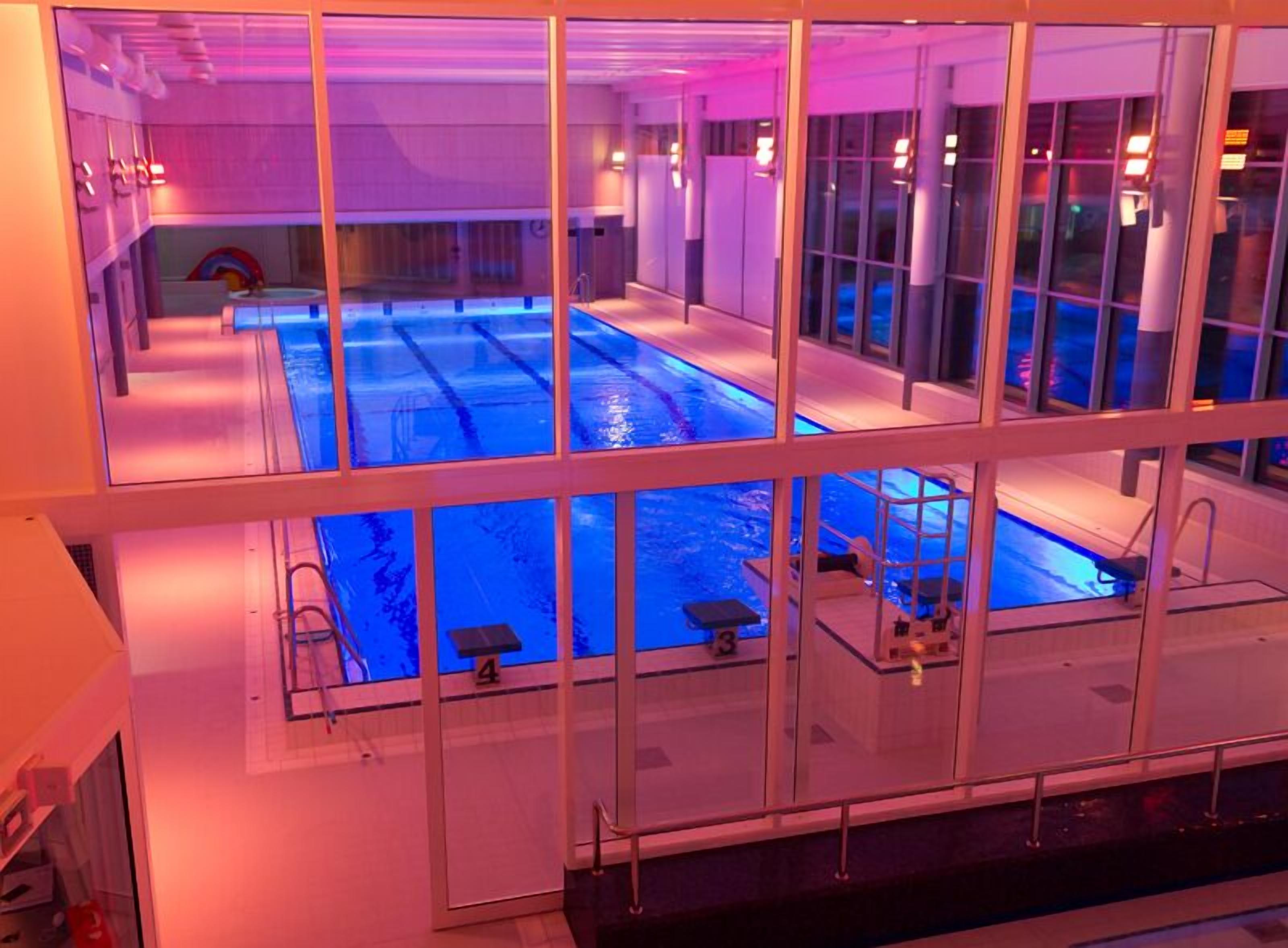 A large indoor swimming pool with multiple lanes and a diving board, surrounded by glass walls.