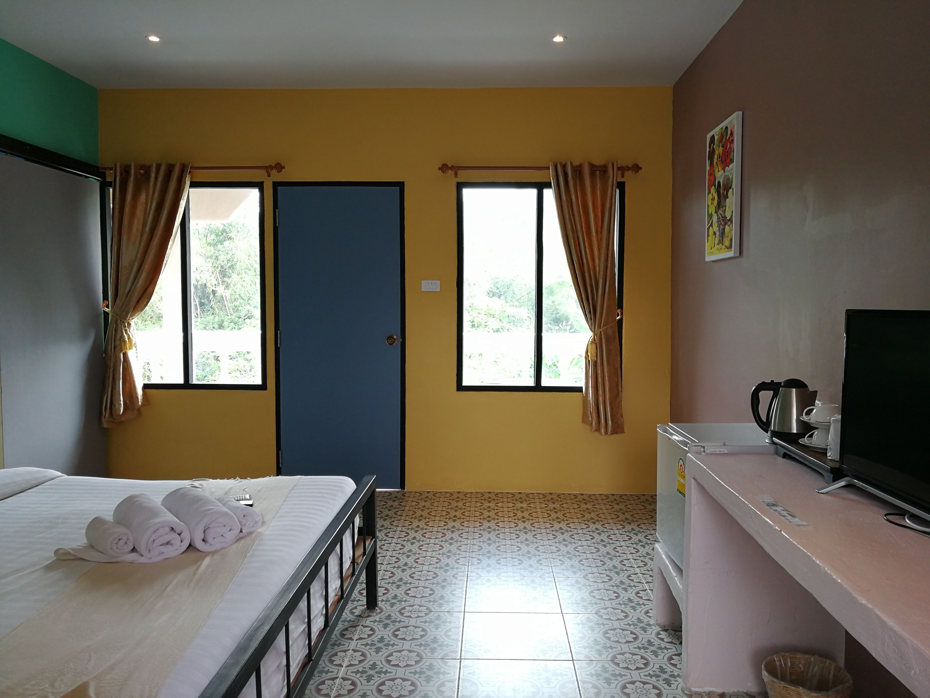 A hotel room with a bed, a desk with a kettle, and two windows with curtains.