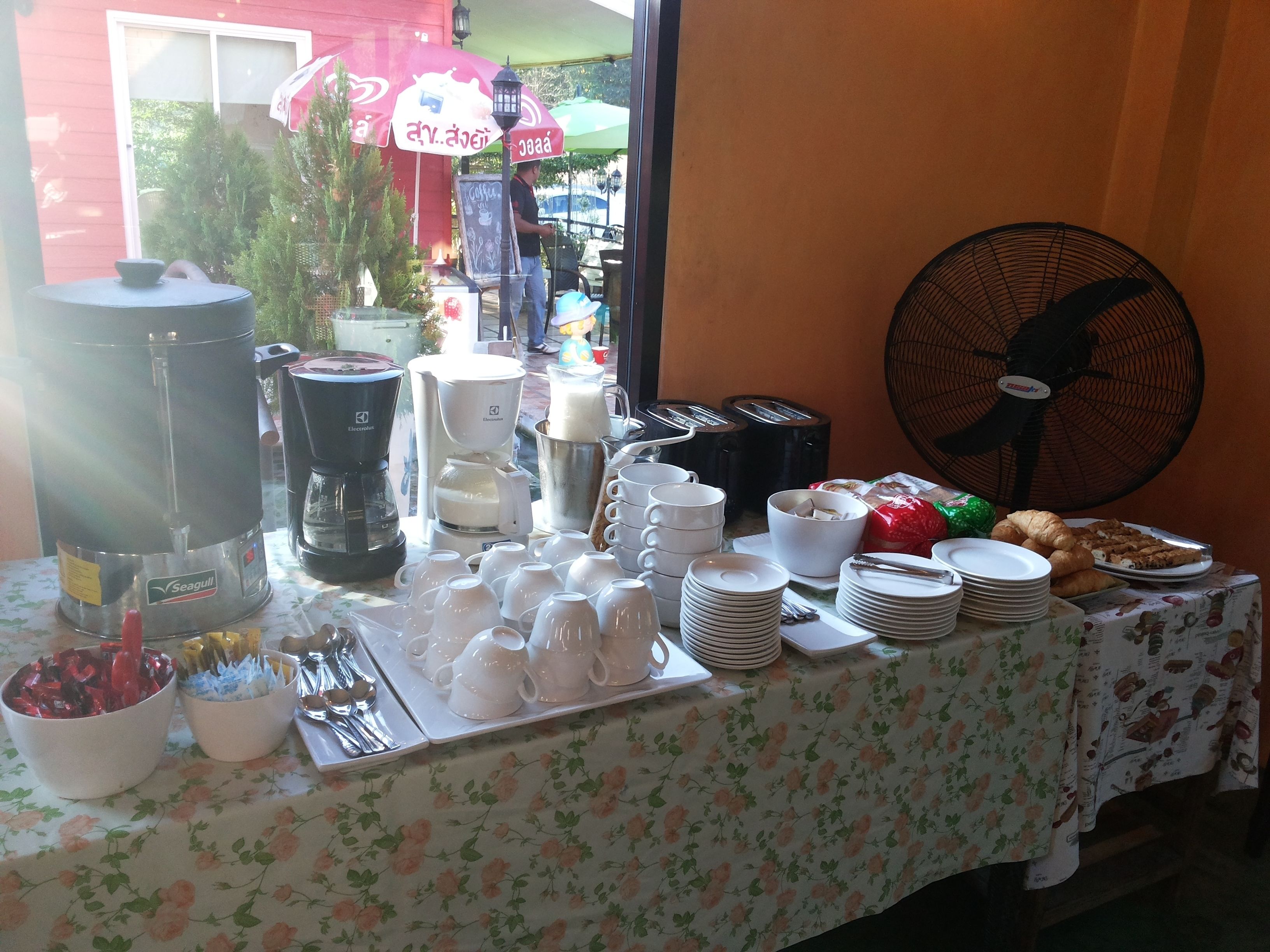 A breakfast buffet setup with various food items, coffee machines, and utensils on a table with a floral tablecloth.
