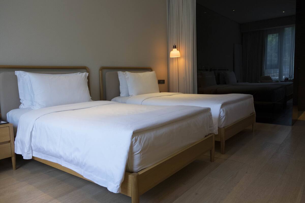 Quarto de hotel com duas camas de solteiro, uma mesinha de cabeceira, um abajur e uma janela com cortinas.
