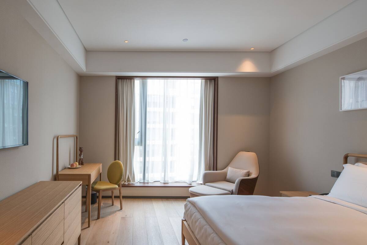 Quarto de hotel com cama, escrivaninha, cadeira e janela com cortinas.