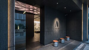 A modern hotel entrance with a dark facade, a glass door, and three wooden stools outside.