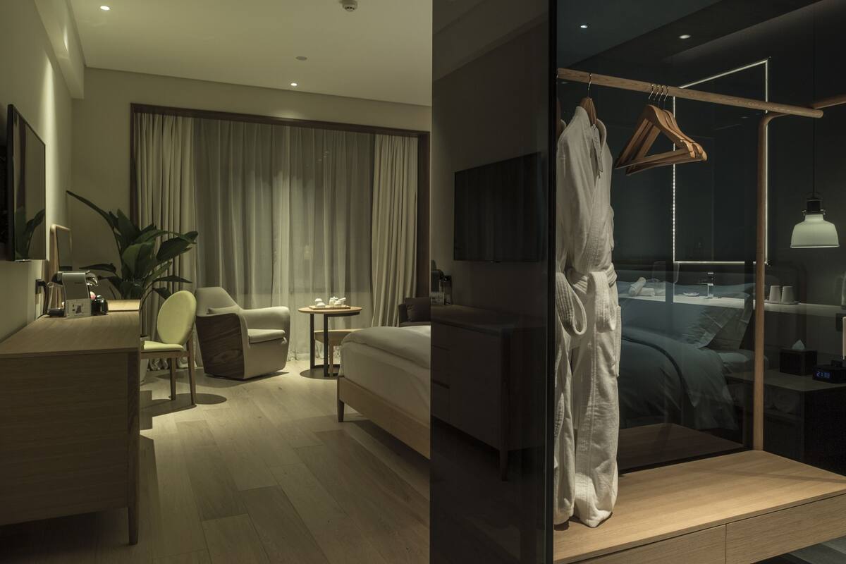 Quarto de hotel moderno com uma cama grande, uma escrivaninha com cadeira, uma mesinha, uma planta em vaso e um guarda-roupa com espelho.