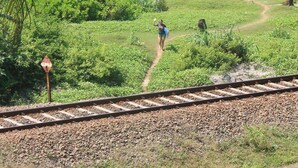 Zona verde con una vía de tren, una persona caminando y vista al océano.