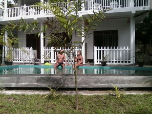A swimming pool with two people sitting on the edge, surrounded by a white fence and potted plants. - Asanka Surf House (Ahangama)