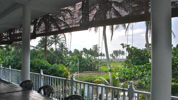 A covered outdoor area with a wooden deck, white railing, and a view of palm trees and greenery.