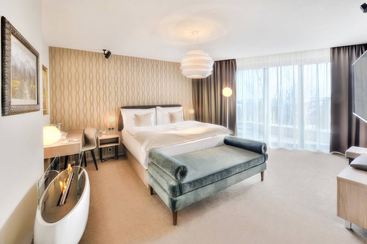 Een hotelkamer met een bed, nachtkastje, lamp en uitzicht op de stad door grote ramen.