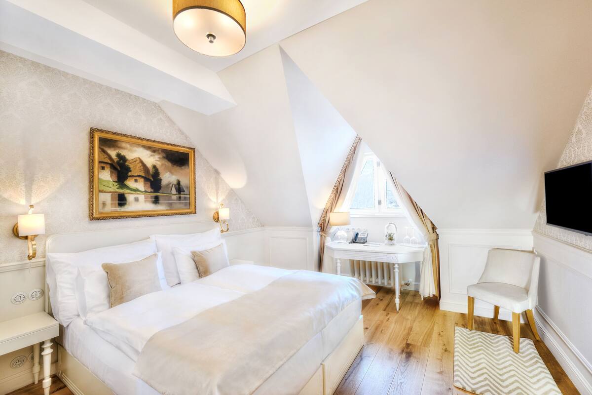 Een slaapkamer met een groot bed, een nachtkastje, een klein bureau met stoel, een raam en een aan de muur bevestigde televisie.